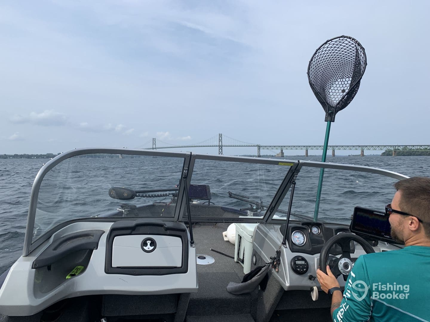 Interior view of a fishing boat with a captain at the helm, navigating through a scenic waterway with a bridge in the background.