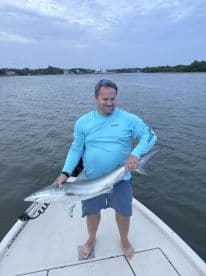 Angler proudly displaying a caught shark while standing on the bow of a boat in calm waters.