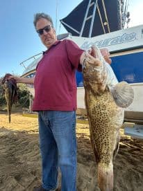 An angler proudly displaying a large Grouper caught during a fishing trip, with a boat in the background.