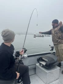 A young angler is actively fishing from a boat in foggy conditions, while an adult demonstrates casting techniques.
