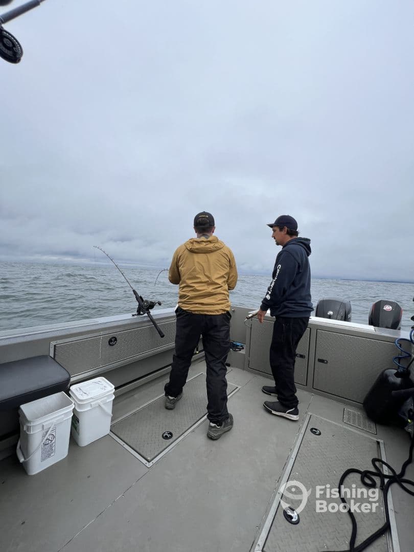 Two anglers actively fishing on a boat, with one preparing to reel in a catch while the other assists, set against a cloudy backdrop.