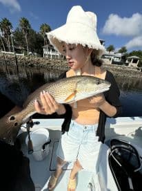 An angler proudly displaying a large Redfish while fishing on a sunny day, with palm trees and a waterfront property in the background.