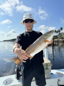 Young angler proudly displaying a Redfish while fishing from a boat in a scenic coastal environment.
