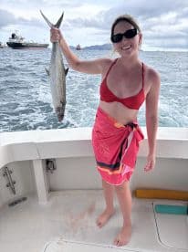 A young woman proudly displaying a large fish while aboard a fishing boat, with a scenic backdrop of cargo ships in the distance.