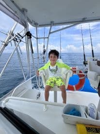 A young boy enjoying his time on a fishing charter, seated in the helm area of the boat, surrounded by fishing gear and a scenic ocean backdrop.