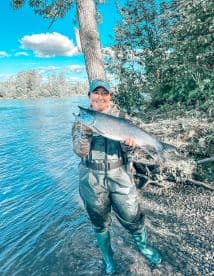 Angler proudly displaying a large Salmon while standing in a river, surrounded by lush greenery.
