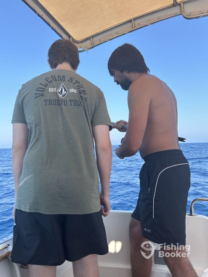 Two anglers engaged in fishing on a boat, with one assisting the other in reeling in a catch in open water.