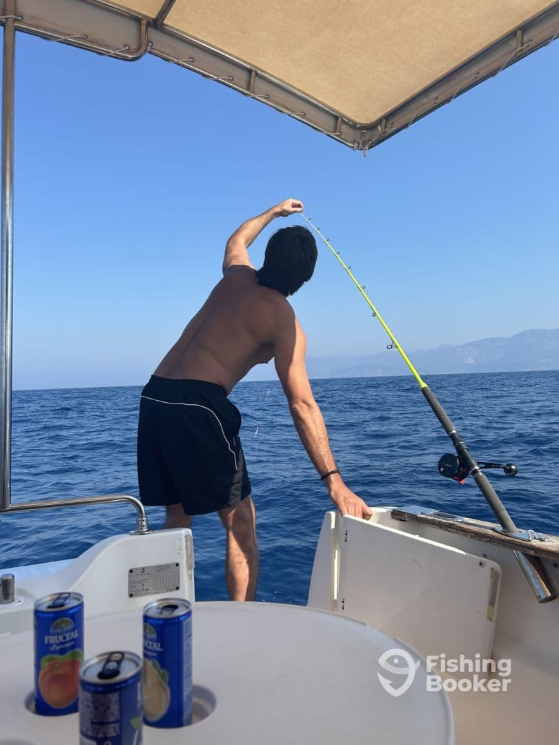 A shirtless angler reeling in a catch while enjoying a day of fishing on a boat in clear blue waters.