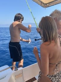 A group of anglers enjoying a fishing trip on a boat, with one person reeling in a small fish in clear blue waters.