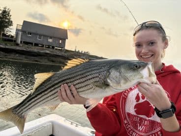 A young woman proudly displaying a large Striped Bass during a sunset fishing trip in a scenic coastal setting.