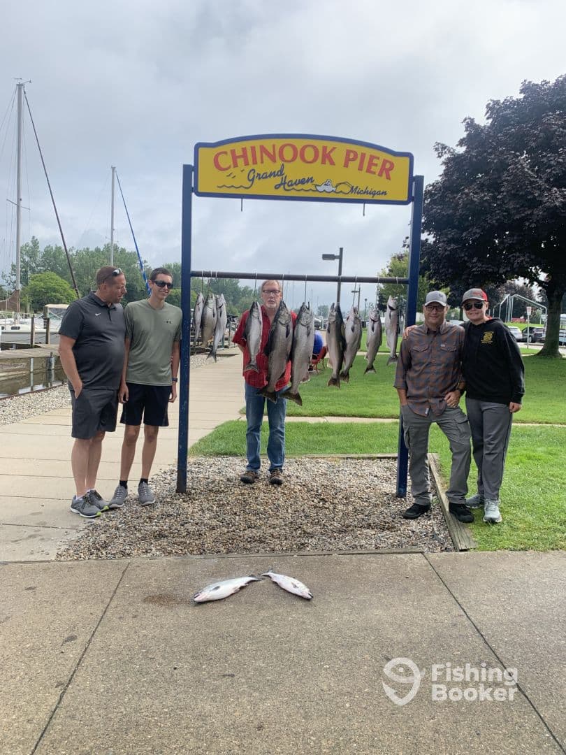 Group of anglers proudly displaying their catch of Chinook Salmon at Chinook Pier in Grand Haven, Michigan, with additional fish laid out on the ground.
