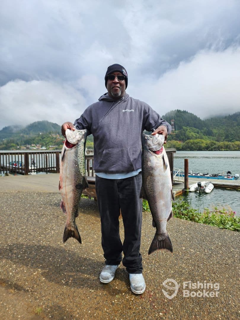 An angler proudly displaying two large Salmon at a scenic dock, showcasing a successful fishing trip.
