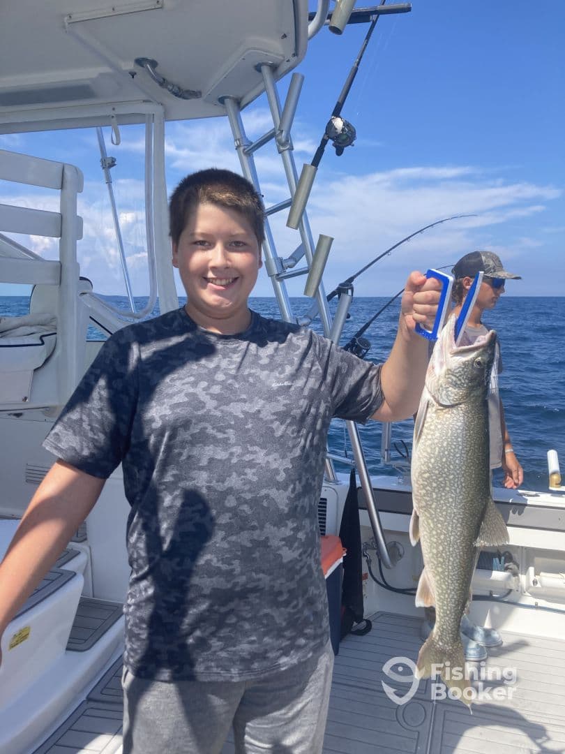 A young angler proudly displaying a large Lake Trout while fishing on a boat, enjoying a sunny day on the water.