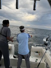 Two anglers actively fishing from a boat, casting lines into the open water under a cloudy sky.