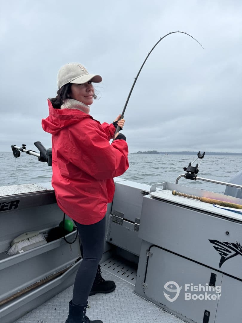 A female angler in a red jacket is actively reeling in a fish while fishing on a cloudy day, showcasing the excitement of the catch.
