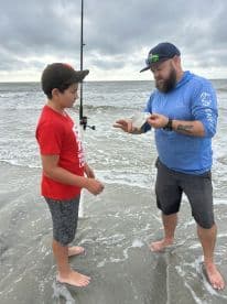 A young angler and an adult are engaged in fishing at the beach, with the adult showing a small fish caught in the surf.