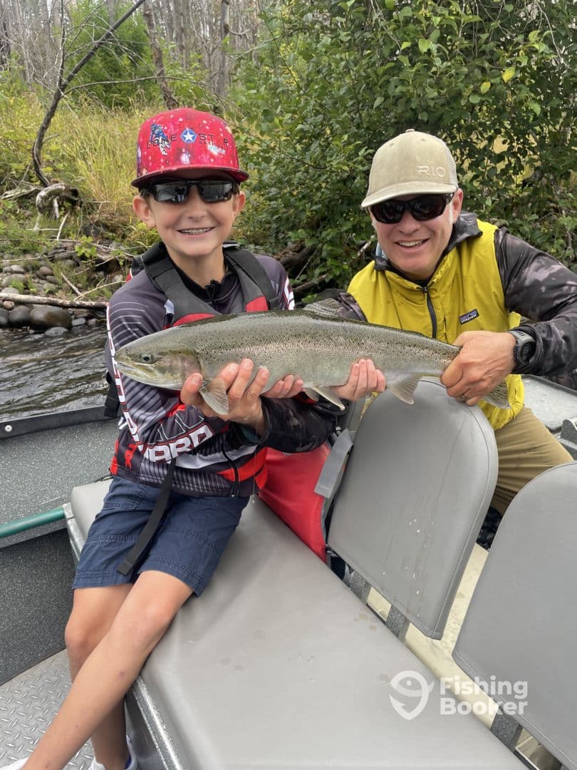 A young angler proudly displaying a large trout while fishing with an adult on a scenic river.