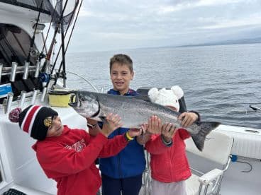 Three young anglers proudly displaying a large Salmon on a fishing boat, showcasing a successful day on the water.