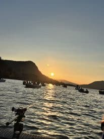 A scenic sunset fishing scene on a lake, with multiple boats in the background and a fishing rod in the foreground.