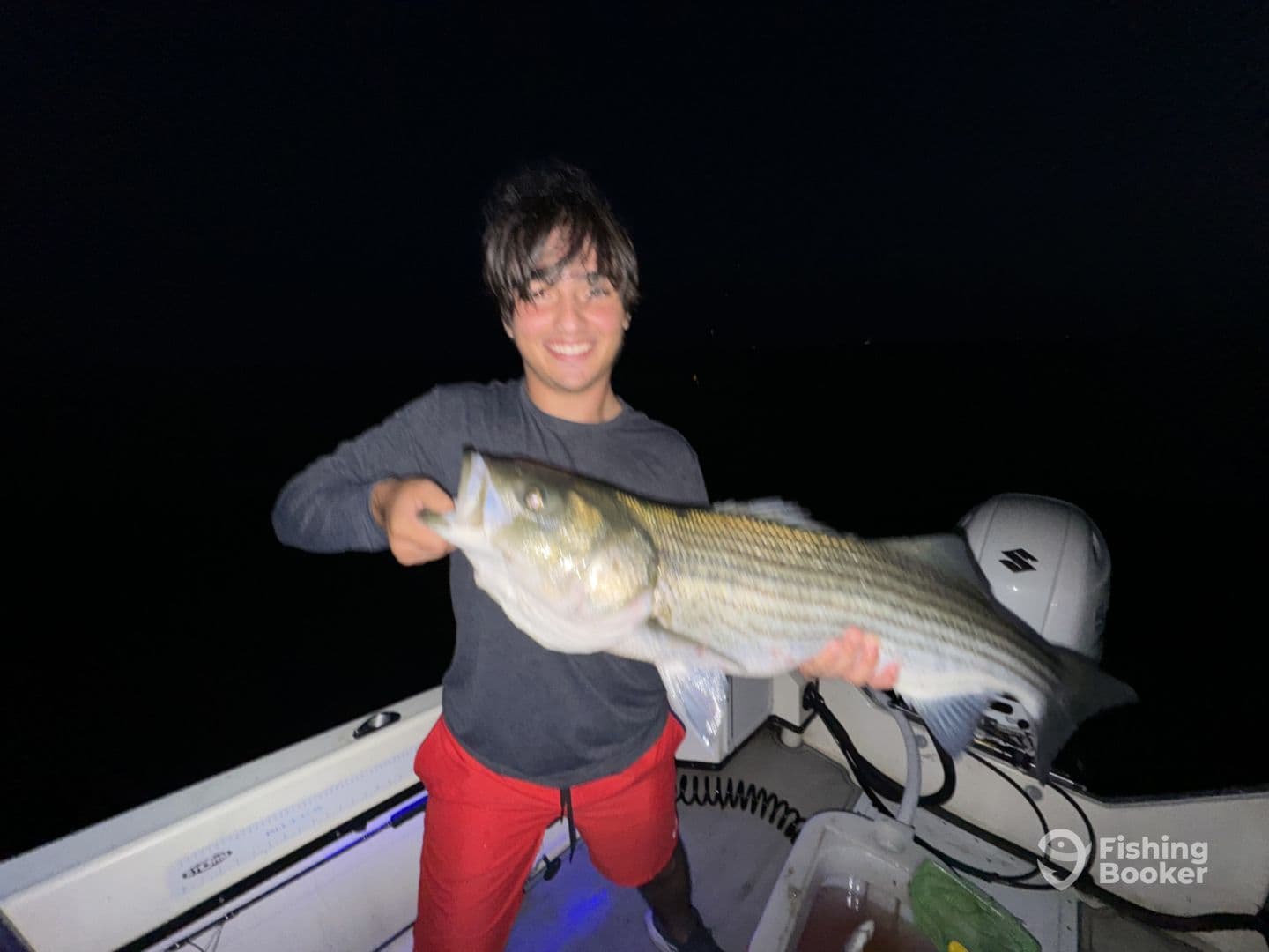 A young angler proudly displaying a large striped bass while fishing at night on a boat.