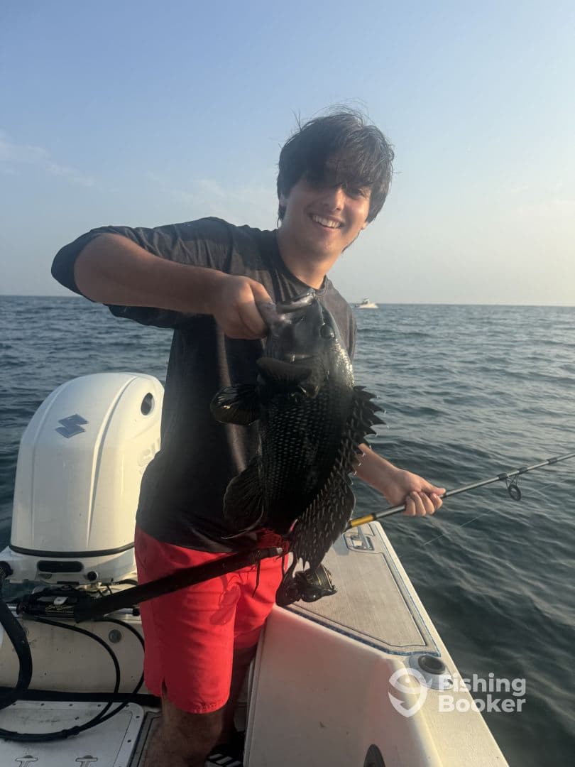 A young angler proudly displaying a Black Sea Bass while fishing on a boat in open waters.