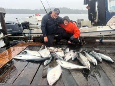 A proud angler and a young boy display their catch of multiple fish, including halibut and salmon, on a rainy day at the dock.