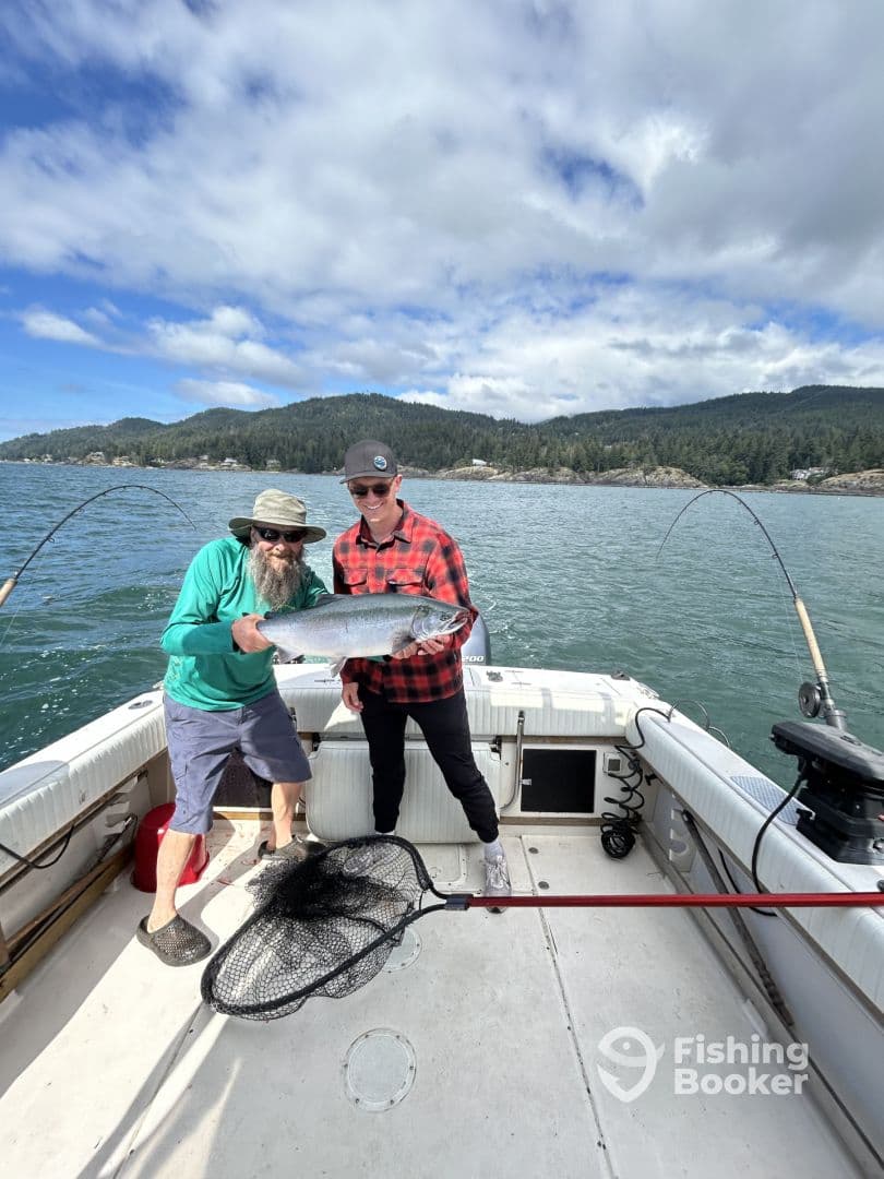 Two anglers proudly displaying a large Salmon on a fishing boat in a scenic coastal environment.