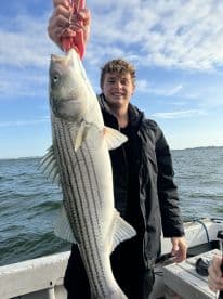 A young angler proudly displaying a large Striped Bass while fishing on a boat, showcasing a successful day on the water.