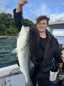 A young angler proudly displaying a large Striped Bass while fishing on a boat, surrounded by a scenic waterway.