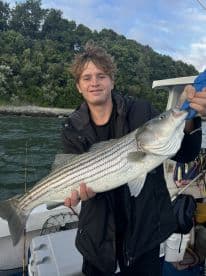 Young angler proudly displaying a large striped bass while fishing on a boat, surrounded by a scenic backdrop of greenery.