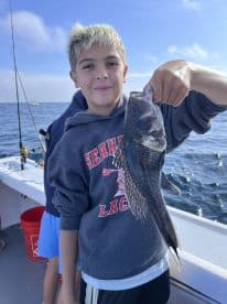 A young angler proudly displaying a Black Sea Bass while fishing on a boat, showcasing a successful day on the water.