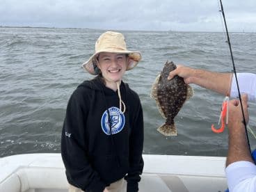 A young angler smiles proudly while holding a Flounder during a fishing trip on a boat, showcasing the excitement of catching fish in a coastal environment.