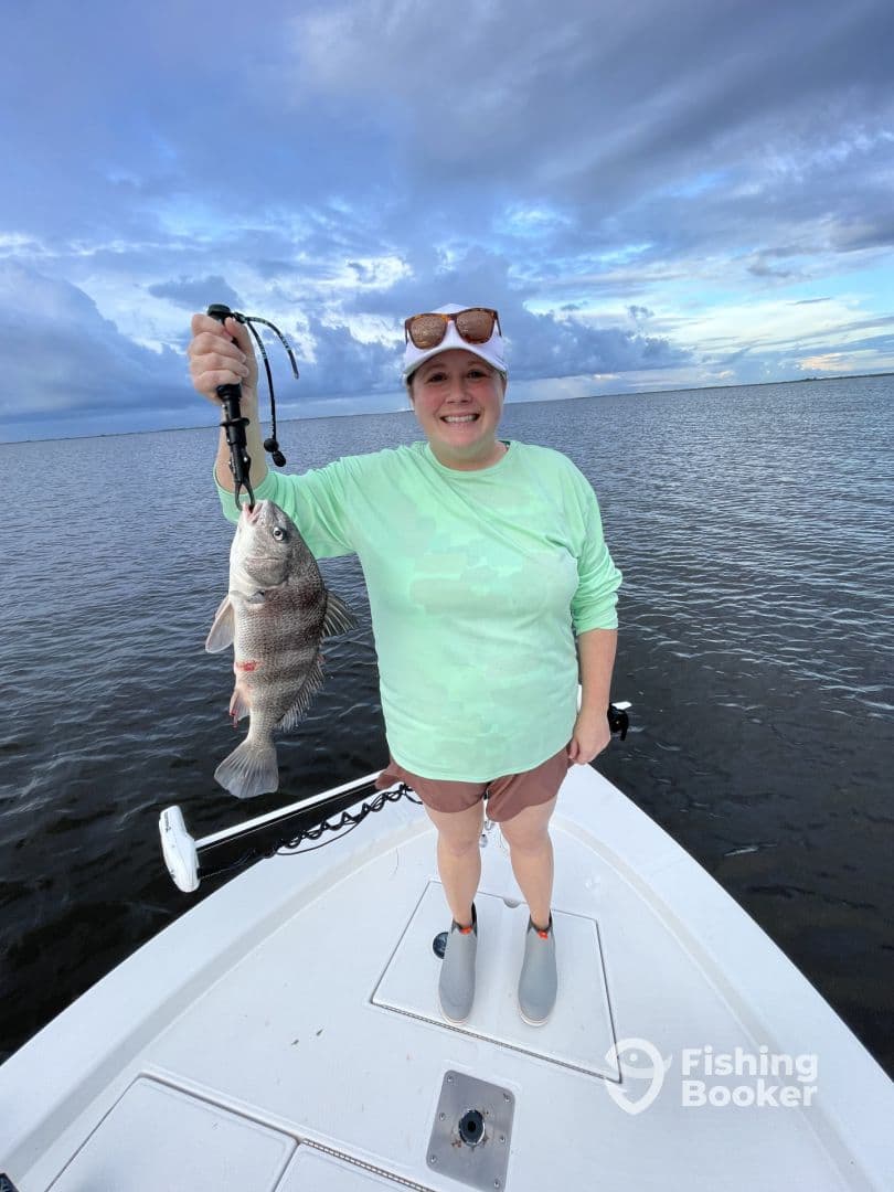 Angler proudly displaying a large Black Drum while standing on the bow of a boat in calm waters.