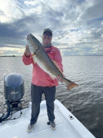 Angler proudly displaying a large Redfish while standing on a boat in a scenic fishing environment.