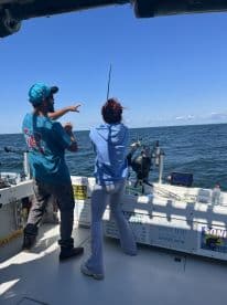 Two anglers engaged in fishing on a boat, with one reeling in a catch while the other points towards the water, showcasing an active fishing experience.