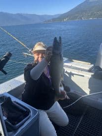 An angler proudly displaying a large Lake Trout while fishing on a scenic lake, surrounded by mountains.