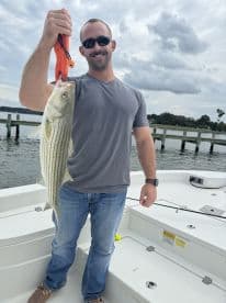 An angler proudly displaying a striped bass caught during a fishing trip, with a scenic backdrop of the water and dock.