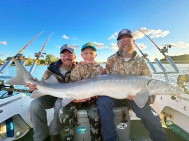 A young boy proudly holds a large Sturgeon while fishing with two adults on a boat, showcasing a successful day on the water.