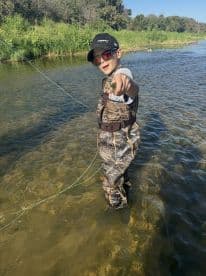 A young angler proudly displaying a small catch while standing in a clear stream, showcasing a fun day of fishing in a natural setting.