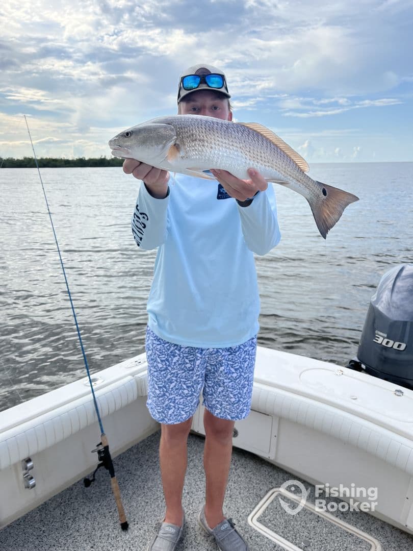 Young angler proudly displaying a Redfish while fishing on a boat in a scenic coastal environment.