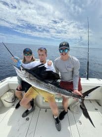 Three anglers proudly displaying a large Sailfish aboard a fishing boat in open waters, showcasing a successful deep-sea fishing trip.
