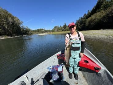 An adult angler proudly displaying a large salmon while fishing from a small boat in a serene river setting, surrounded by lush greenery.