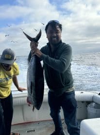 An angler proudly displaying a large tuna on a fishing charter, with a scenic ocean backdrop and another crew member in the frame.