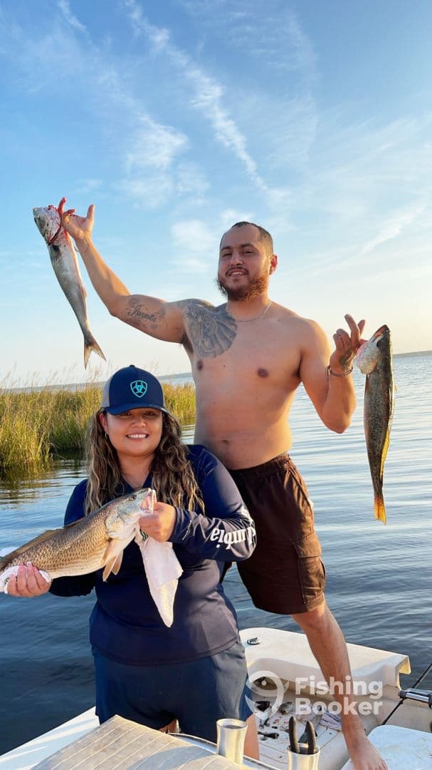 Two anglers proudly displaying their catches, a large Speckled Trout and a Redfish, while enjoying a fishing trip in a scenic coastal environment.