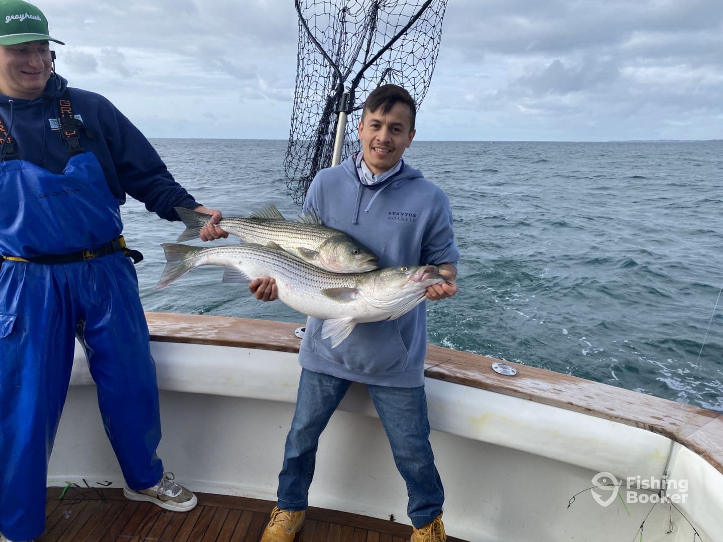 Two anglers proudly displaying their catch of striped bass aboard a fishing boat, with a scenic ocean backdrop.