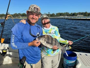 Two anglers proudly displaying a large Black Drum while fishing on a boat in calm waters.