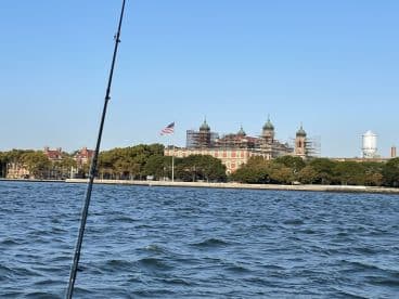 A scenic view of the boat with a fishing rod in the foreground, showcasing the historic Ellis Island in the background, under renovation.