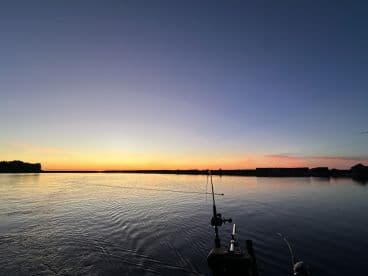A serene sunset view over calm waters, with fishing rods visible, suggesting a peaceful fishing environment.