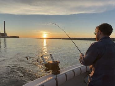 A lone angler is reeling in a fish during a stunning sunset on a calm waterway, showcasing the serene fishing environment.