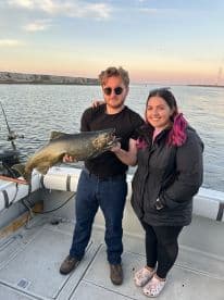 Two anglers proudly displaying a large Lake Trout while fishing on a boat during sunset.